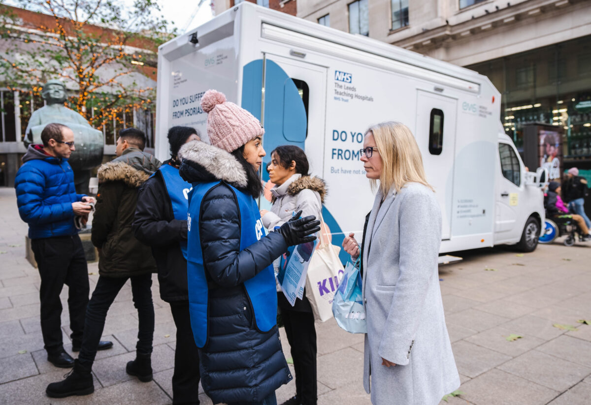 A volunteer in a blue tabard speaking with a member of the public in front of a psoriasis mobile clinical van.