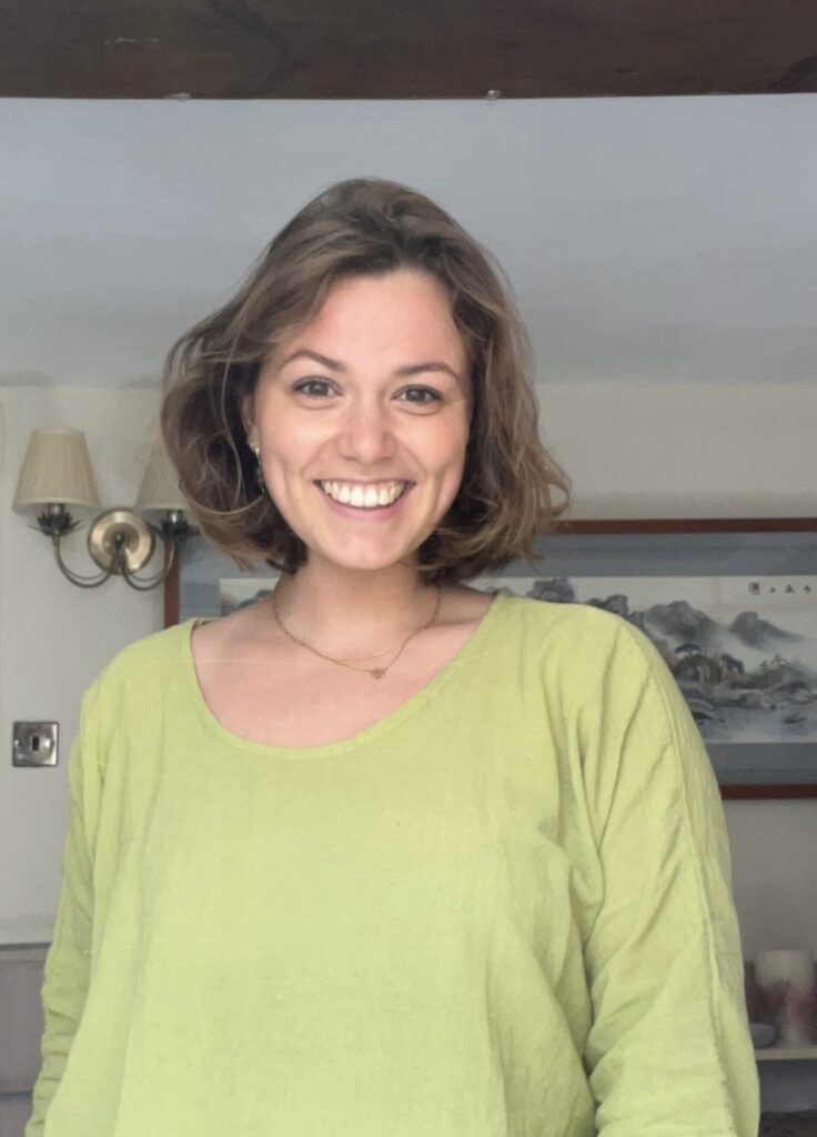 A picture of Charlotte Campbell-Broad from the middle up against a pale background of a room with a picture on the wall behind her.