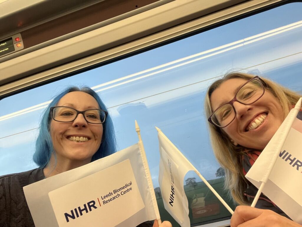 Dr Kerrie Davies (left) with Dr Jane Freeman smiling to camera on a train with small NIHR Leeds Biomedical Research Centre flags.