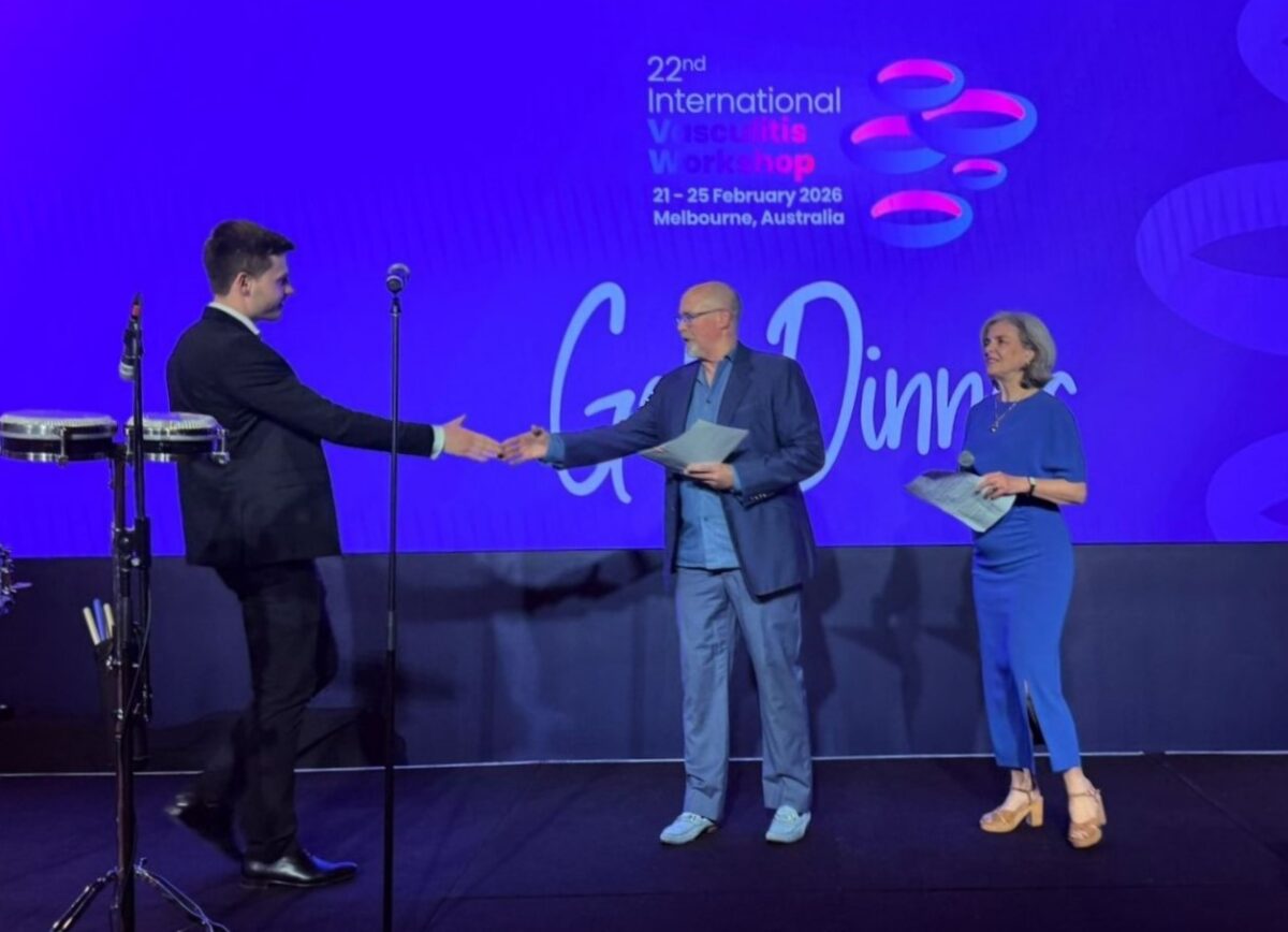 A photo of Dr Michal Zulcinski on stage receiving their award for their research with two presenters on stage in front of a blue background and the name of the conference.