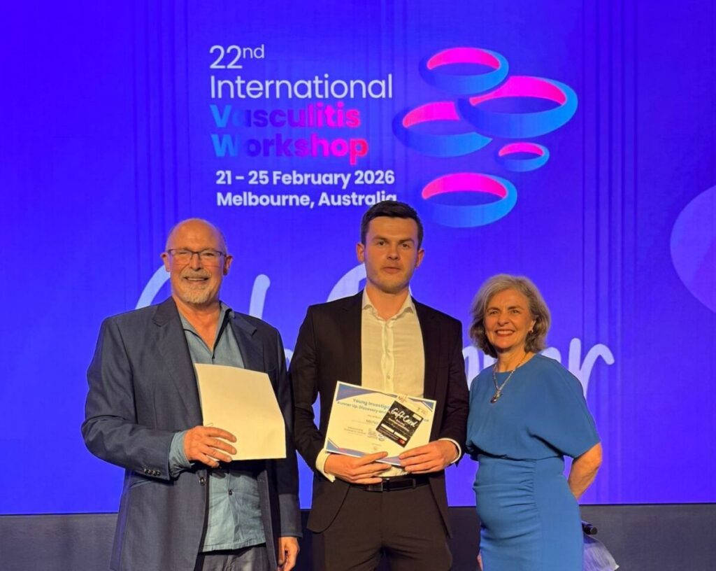 A photo of Dr Michal Zulcinski on stage with their award for their research standing with two presenters one either side on stage in front of a blue background and the name of the conference.